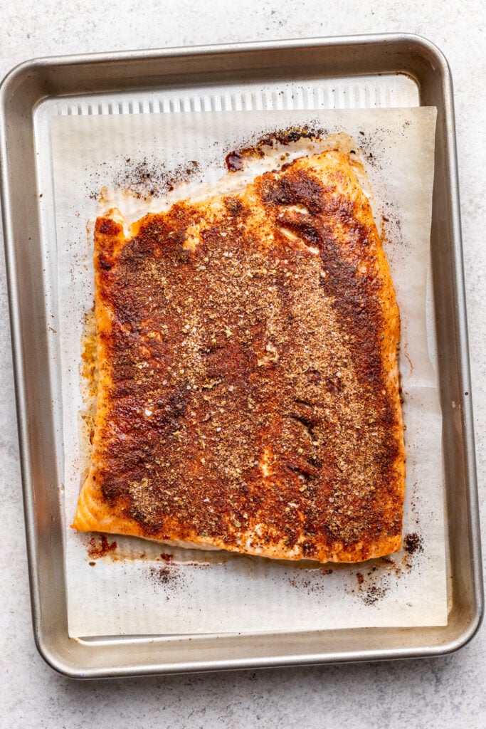 A large, seasoned fillet of salmon sits on a piece of parchment paper atop a baking sheet. The salmon is coated with a dark spice rub and appears ready to be cooked or freshly baked.