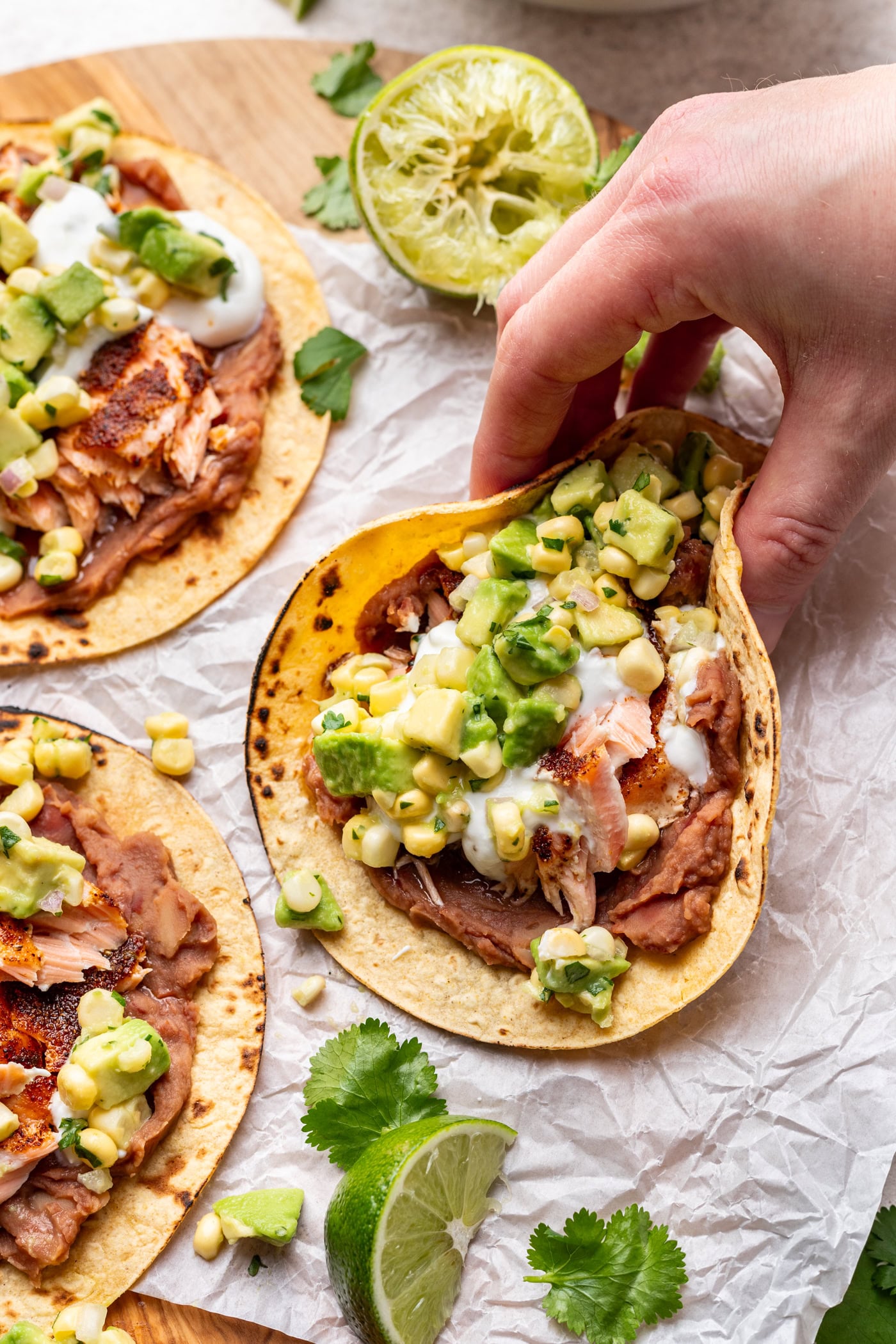 A hand holds a taco filled with grilled fish, refried beans, corn, avocado, and a creamy sauce on a corn tortilla. More tacos, cilantro, and lime wedges are on parchment paper nearby.