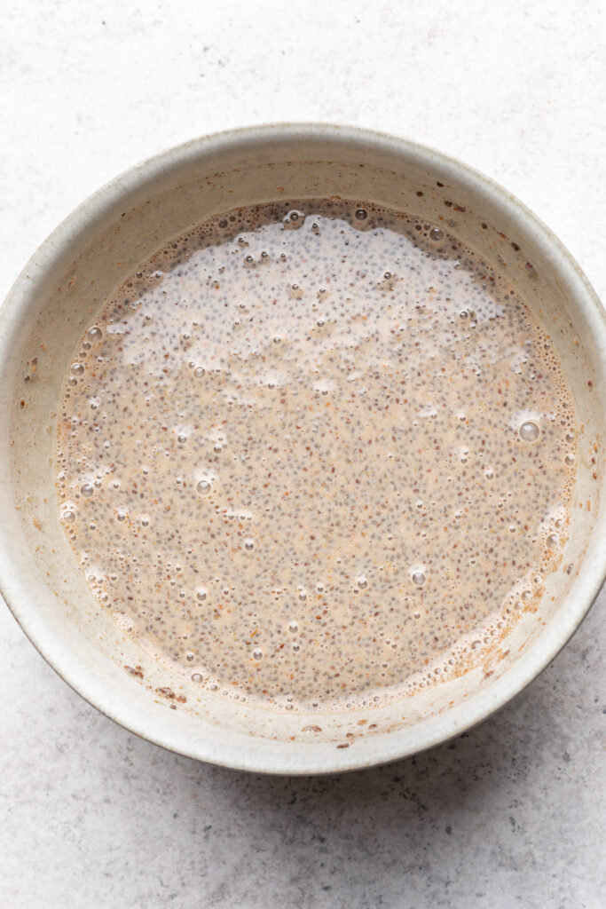 A ceramic bowl filled with a creamy, light brown chia seed mixture, showing hydrated chia seeds suspended in liquid, placed on a light textured surface.