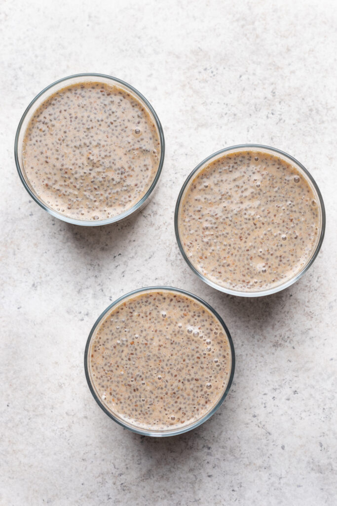 Three glass bowls filled with chia seed pudding arranged in a triangular pattern on a light, textured surface. The pudding has a creamy beige color with visible chia seeds throughout.