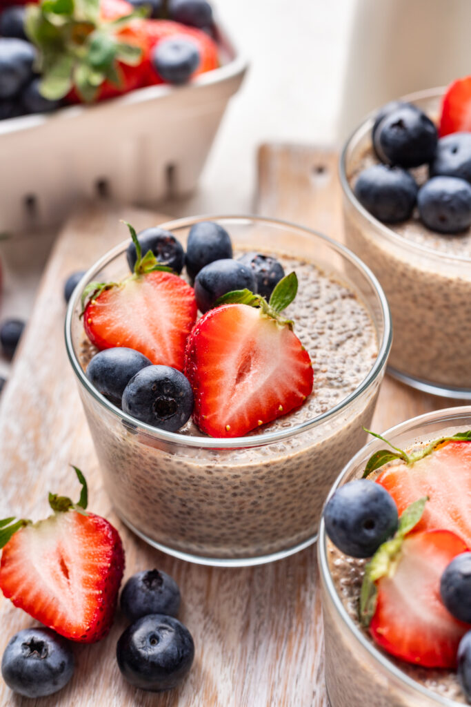 Three glasses of chia pudding topped with fresh strawberries and blueberries are on a wooden surface, with extra berries scattered around and a carton of strawberries in the background.