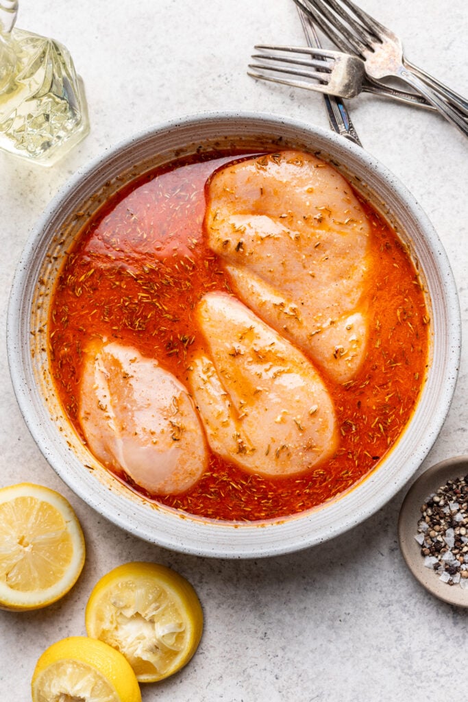 Raw chicken breasts marinating in a bowl of red, seasoned liquid, surrounded by lemon halves, a small dish of pepper, a glass bottle of oil, and several forks on a light surface.