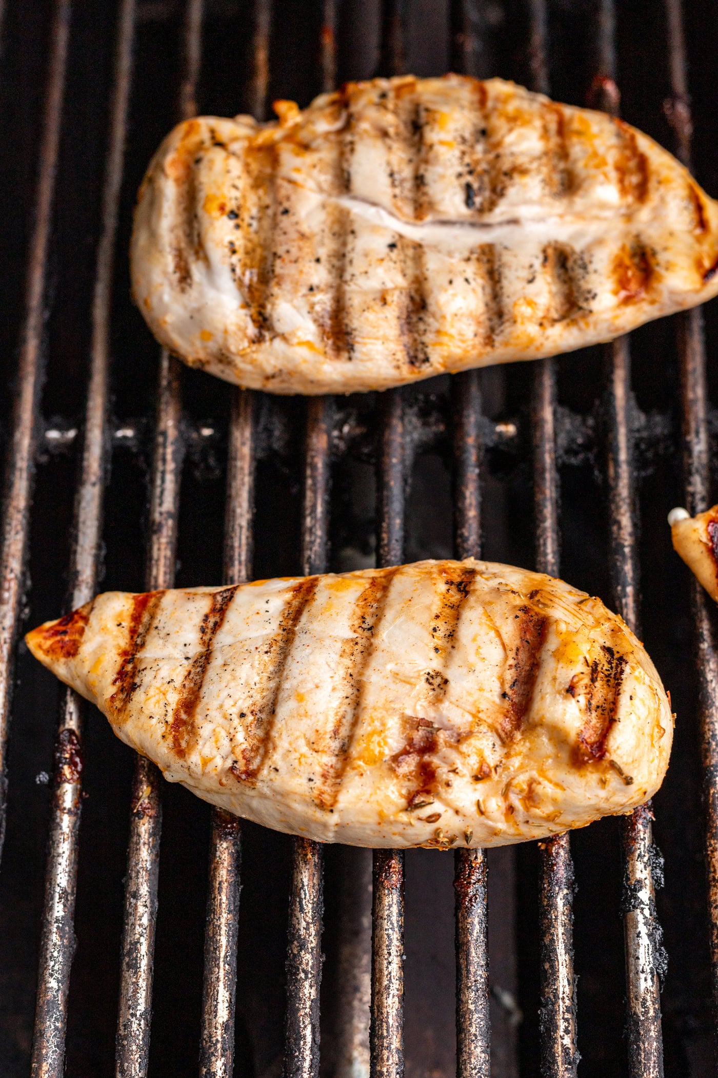 Two pieces of grilled chicken breast with visible grill marks are cooking on a metal grill grate.