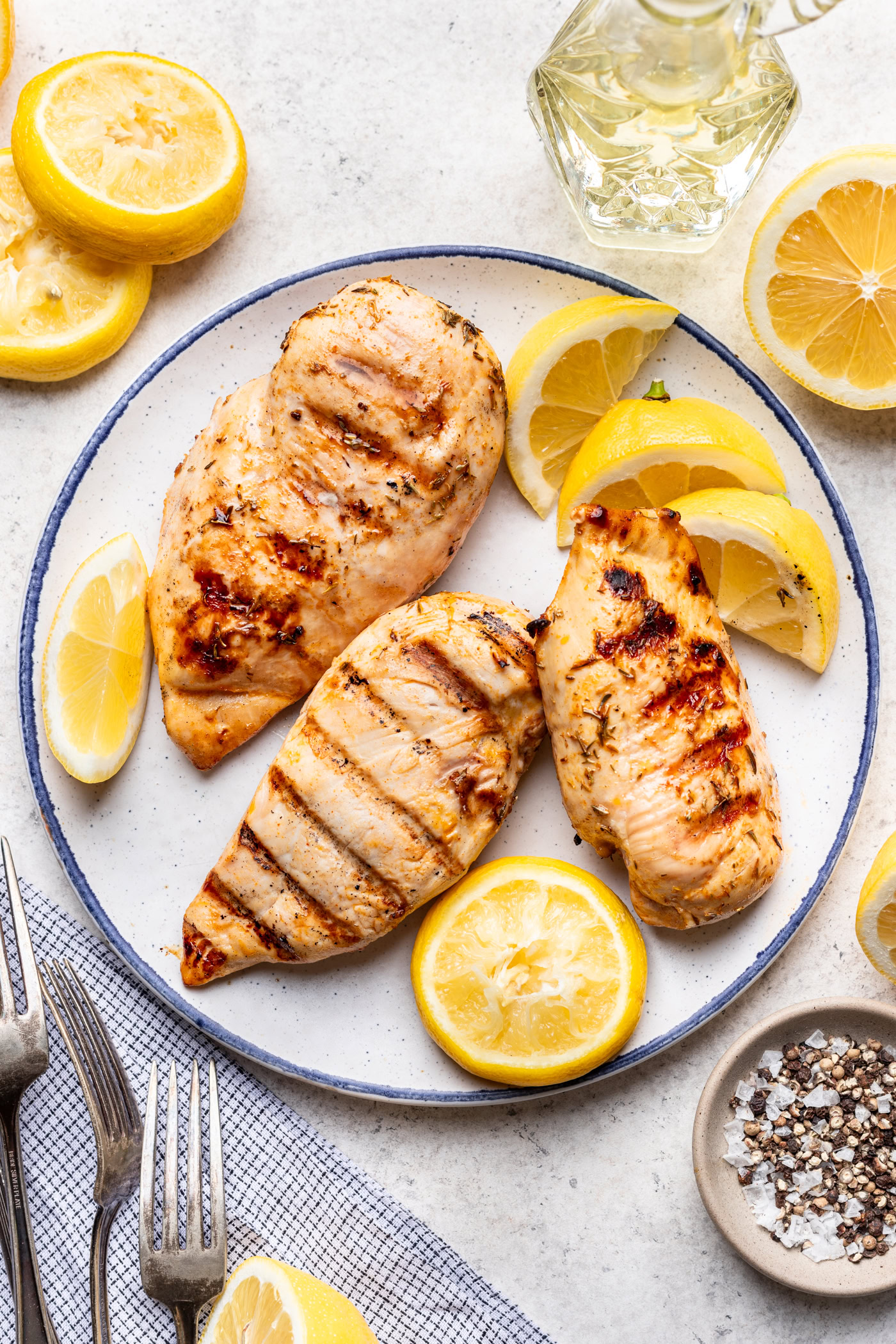 Three grilled chicken breasts on a plate, garnished with lemon slices. Surrounding the plate are more lemon slices, a small bowl of salt and pepper, a glass bottle of oil, and silver forks on a napkin.