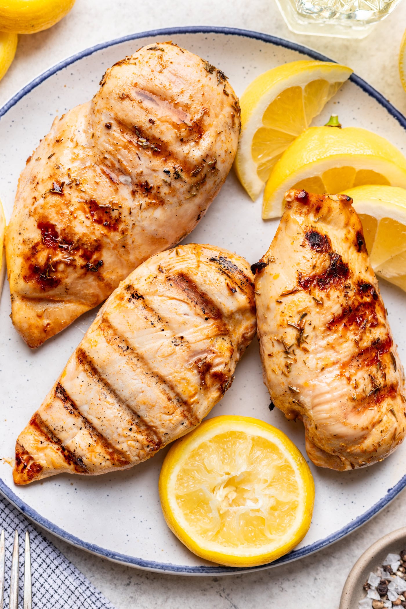 Three grilled chicken breasts on a plate, garnished with fresh lemon wedges. The chicken shows grill marks and is seasoned with herbs. A fork and napkin are visible beside the plate.