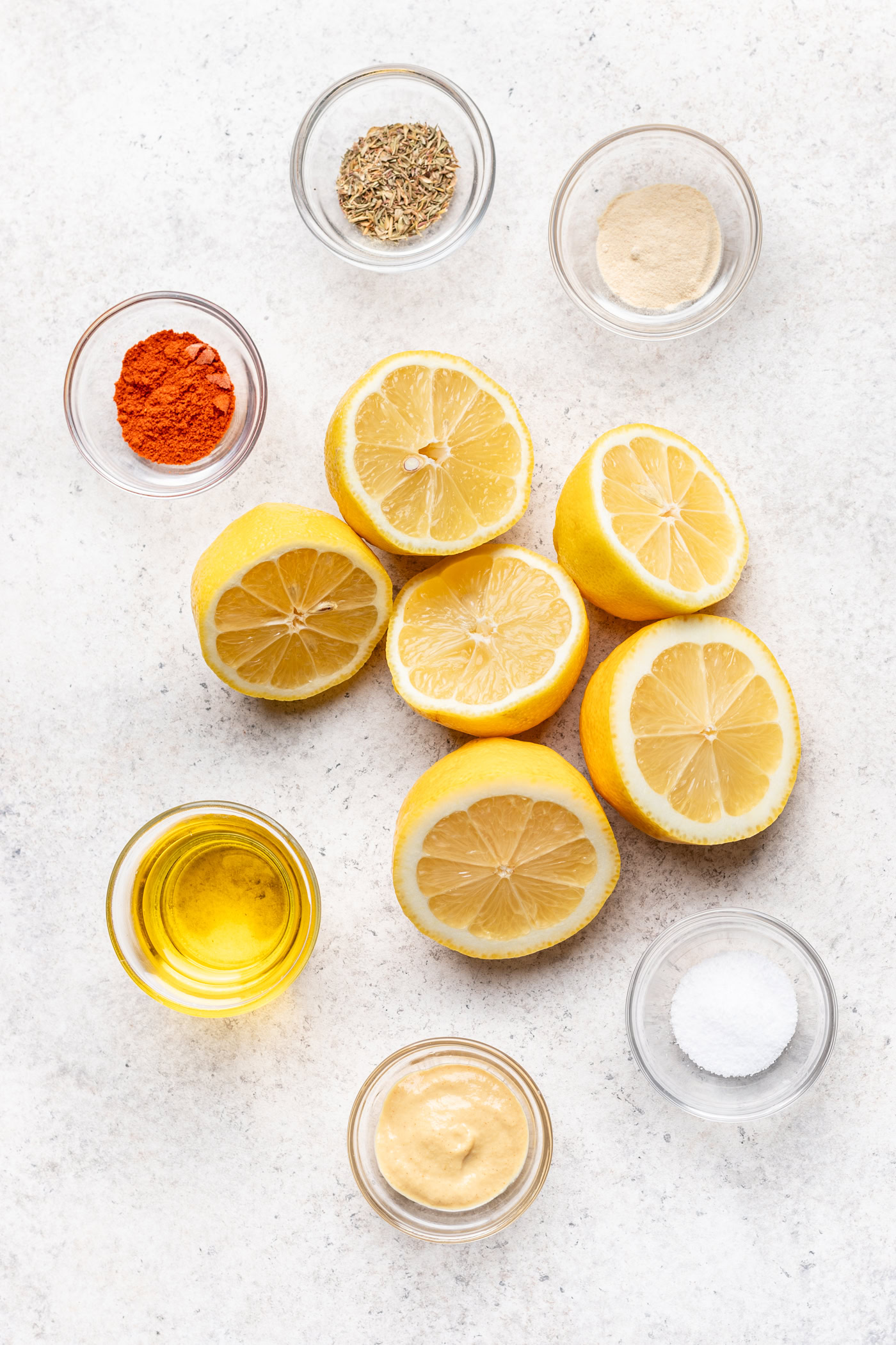 Halved lemons surrounded by small bowls containing olive oil, salt, ground spices, dried herbs, and mustard, all arranged on a light surface.
