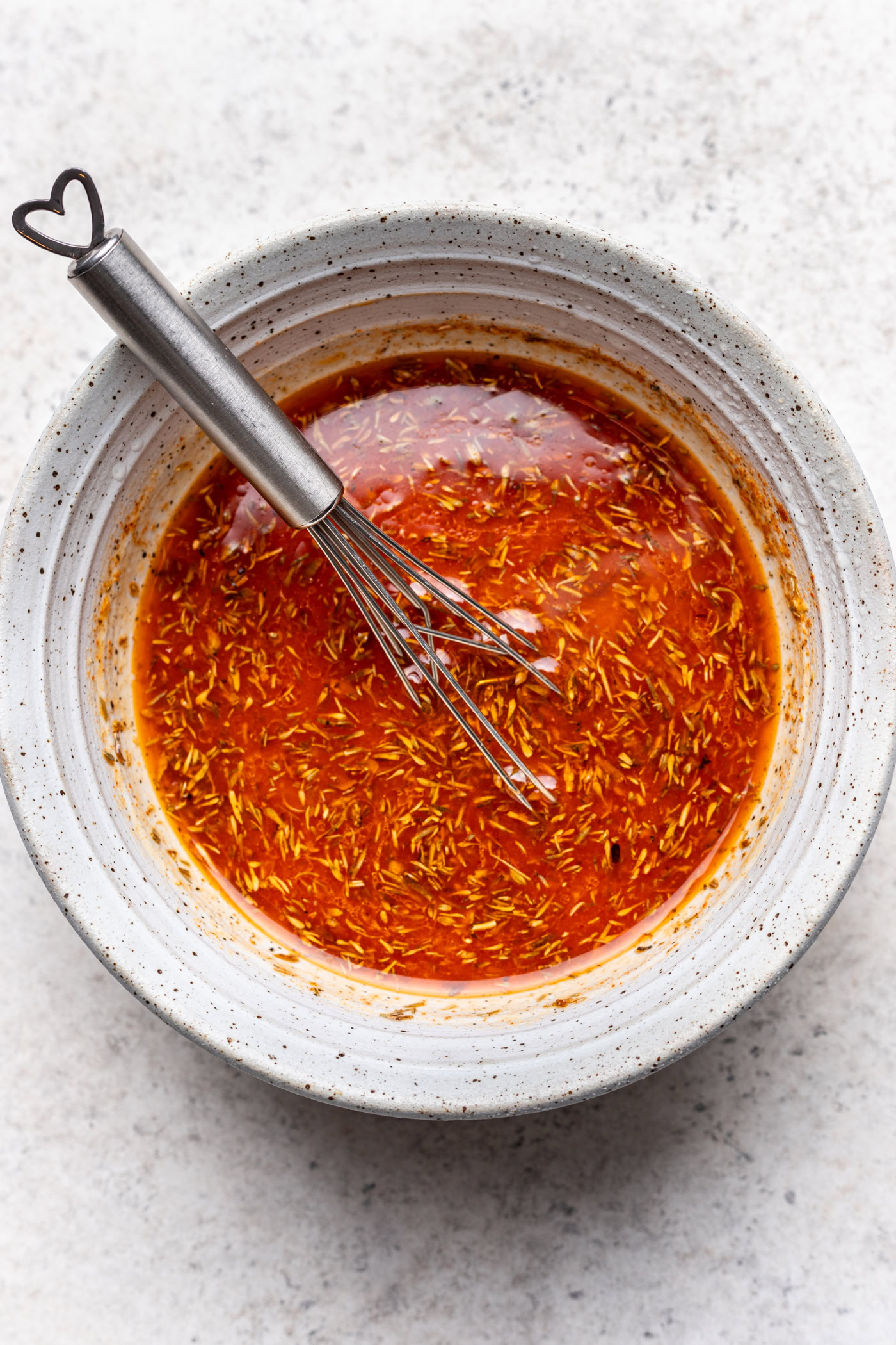 A white speckled bowl filled with a red, oil-based marinade containing visible herbs. A metal whisk with a heart-shaped handle rests inside the bowl. The background is a light, textured surface.