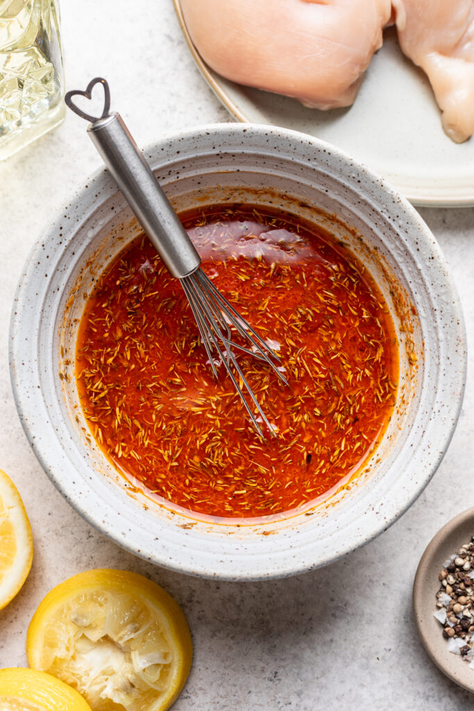 A bowl of red marinade with a whisk inside, surrounded by lemon slices, black pepper, oil, and raw chicken breasts on a plate.