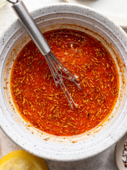 A white bowl filled with red-orange marinade and herbs, with a metal whisk inside. A lemon wedge and part of another dish are visible next to the bowl.