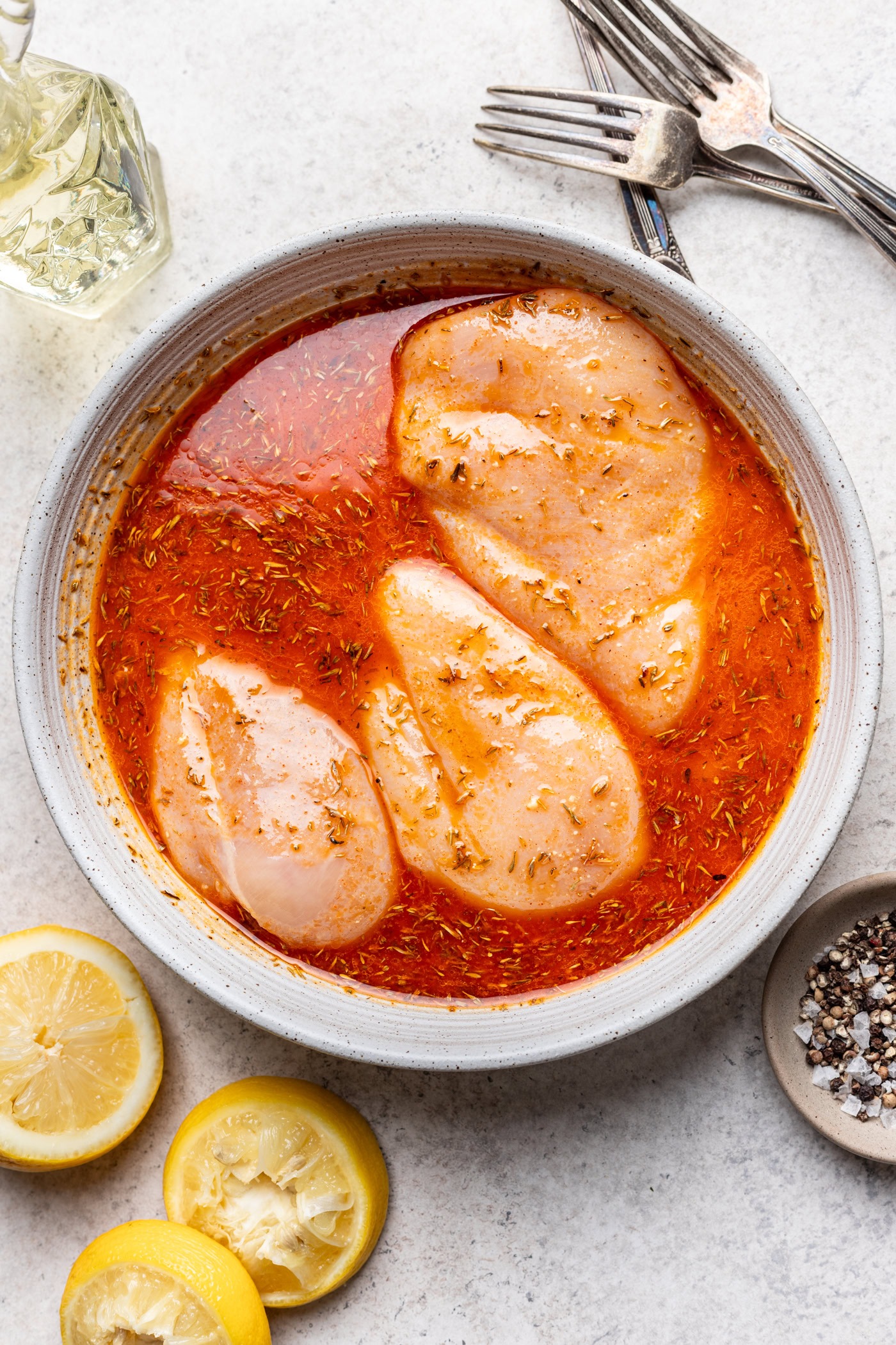 Raw chicken breasts marinating in a red, seasoned liquid in a bowl, surrounded by lemon halves, a bowl of pepper, two forks, and a glass container of oil on a light countertop.