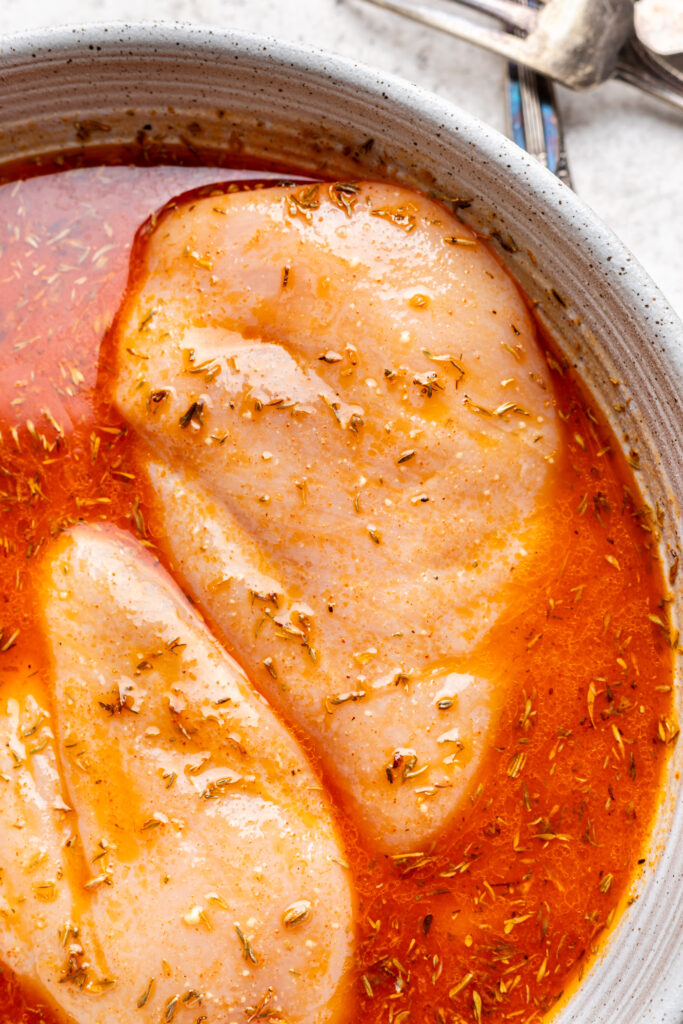 Two seasoned, raw chicken breasts marinating in an orange-red liquid with herbs in a round, white dish. Silver forks are partially visible in the background.