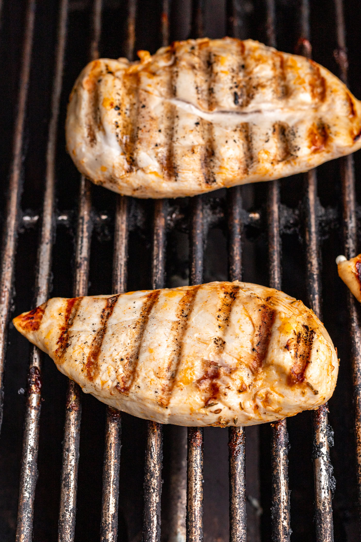 Two pieces of grilled chicken breast with visible grill marks are cooking on a barbecue grill. The chicken is lightly browned and the grill grates are blackened from use.