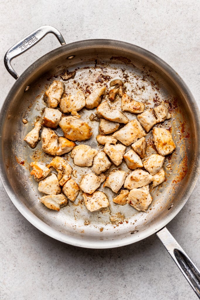 Cooked, browned pieces of chicken breast are scattered in a stainless steel skillet with some seasoning and slight charring visible on the pan.