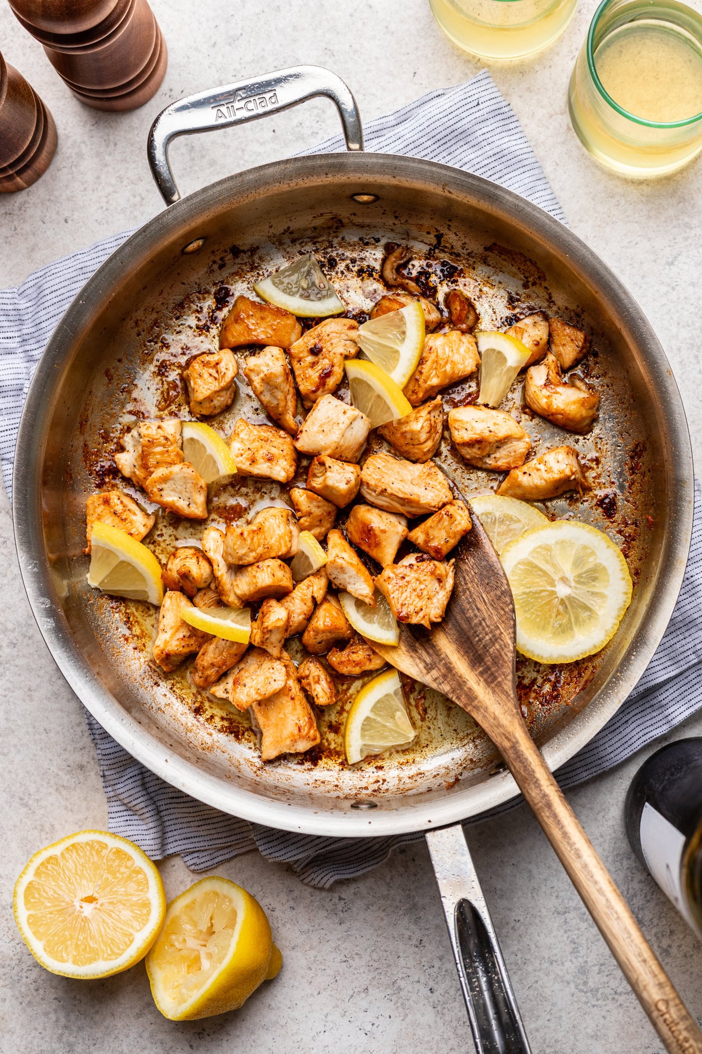 A stainless steel pan with cooked, seasoned chicken pieces and lemon wedges. A wooden spatula rests in the pan. A halved lemon, a striped towel, and drinks are placed nearby on a light countertop.