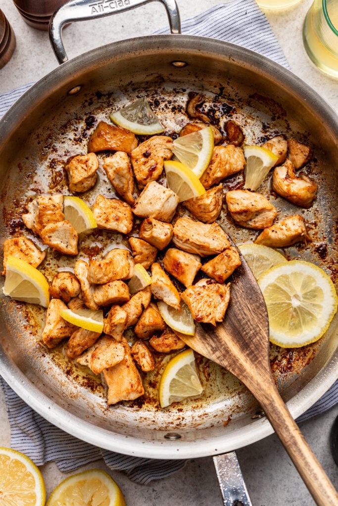 A stainless steel pan with cooked, browned chicken pieces and lemon wedges, stirred with a wooden spoon, sits on a striped cloth with lemon slices nearby.